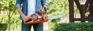 Unrecognizable craftsman standing with cordless electric chain saw, while working in summer garden.