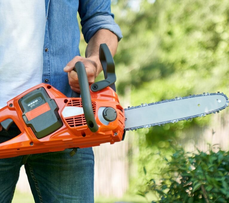 Unrecognizable craftsman standing with cordless electric chain saw, while working in summer garden.