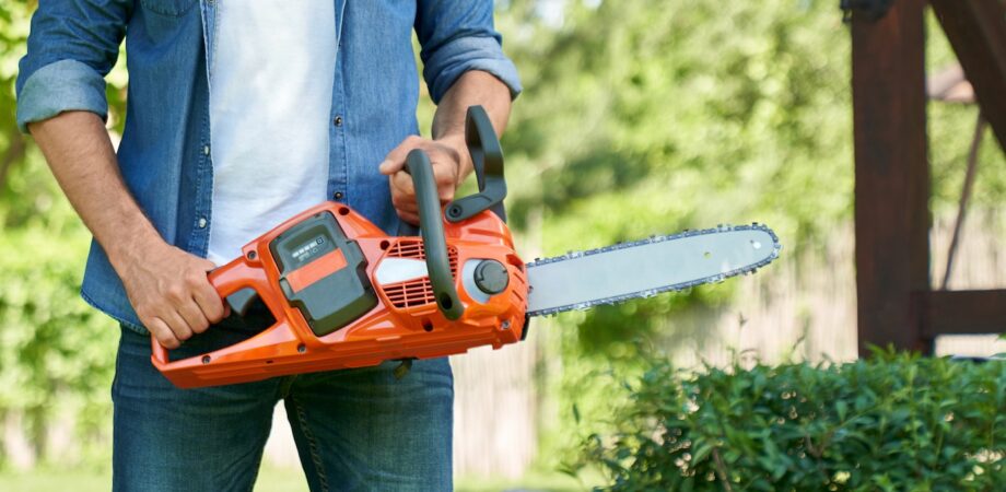 Unrecognizable craftsman standing with cordless electric chain saw, while working in summer garden.